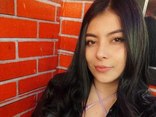 SophiaRamirez has long black hair, brown eyes, and light skin, wearing a black jacket with a pink strappy top, smiling slightly with no makeup, looking cute.