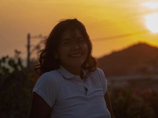 LailaAsher with dark wavy hair, white polo shirt, smiling at sunset, light brown skin, soft evening glow, small nose, casual look, relaxed posture, gentle expression, subtle shadows.