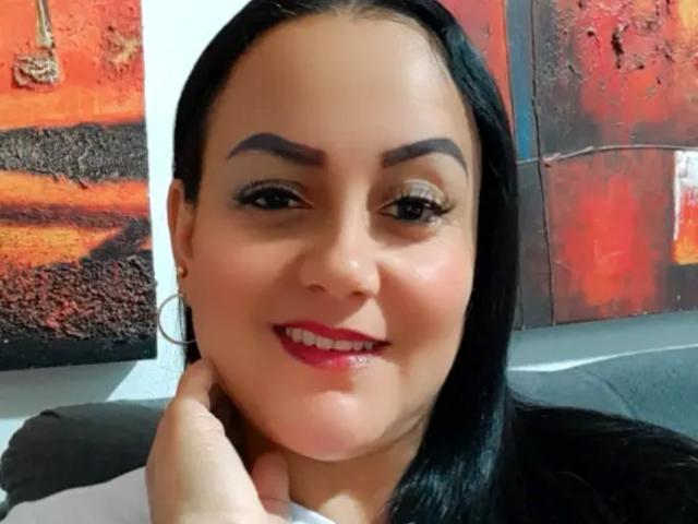 DivaCurvy with black hair, thick eyebrows, red lipstick, and white shirt, smiling, holding her chin, wearing hoop earrings, in a selfie.