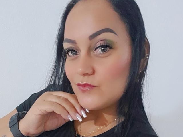 DivaCurvy with long black hair, green and brown eyeshadow, pink lipstick, and white nails, resting her chin on her hand, wearing a black top and a watch.