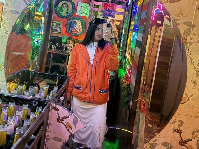 LolaOberle with long black hair, wearing an orange bomber jacket and white dress, holding a phone in a colorful, neon-lit room, standing next to a shelf with jars.