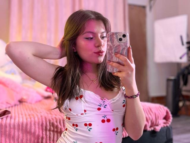 SunBlonde with pink highlights in brown hair, wearing a cherry-print tank top, taking a selfie in a bedroom, pink bed, hand behind head, pink lipstick, cute, attractive.