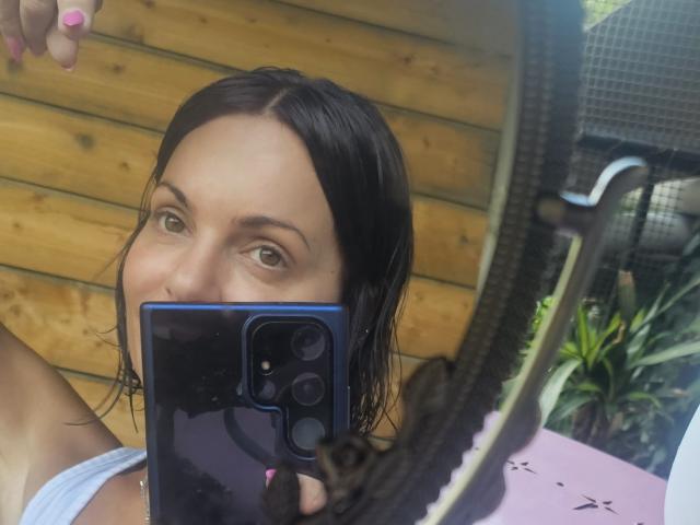 Close-up selfie of JuneLaFrenchie with wet black hair, holding a blue smartphone in front of her face, wearing a white tank top, bike in the background, wooden wall behind.