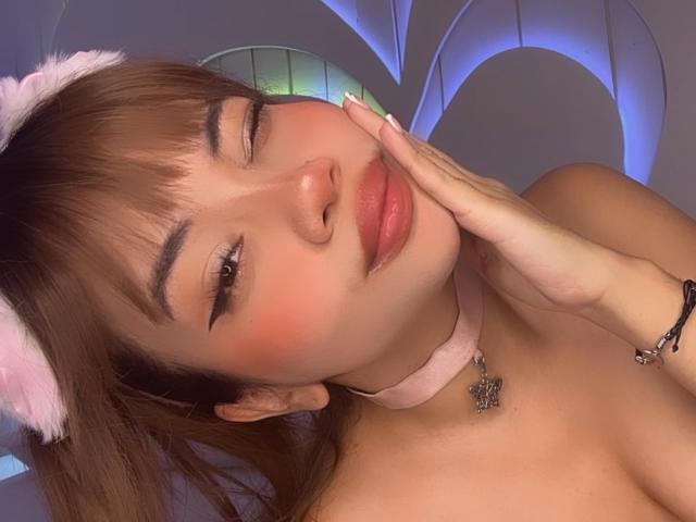 LiliMays with cute pink cat ears, winking, pink blush, and glossy lips, touching her cheek, wearing a pink choker with a butterfly pendant, in a soft, purple-lit setting.