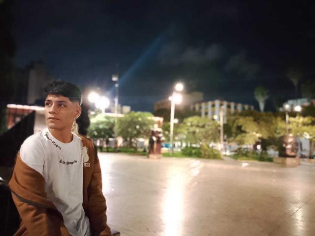 CristoferBlack with short black hair, brown jacket, and white shirt sits on a bench at night, looking to the side with a serious expression. Background is blurred.