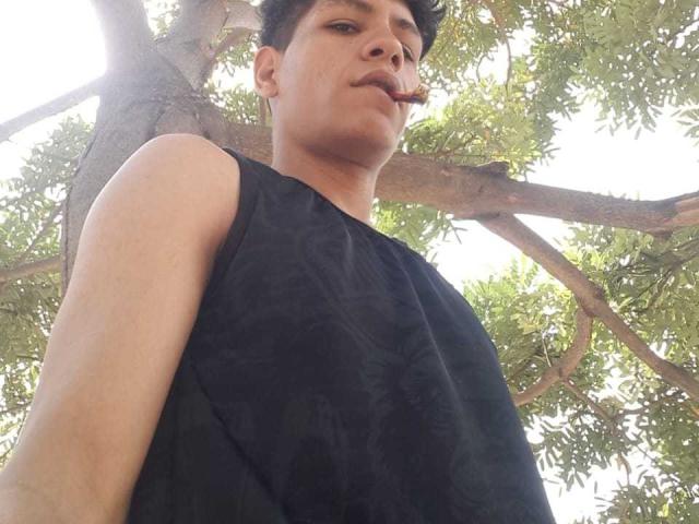 CristoferBlack, wearing a sleeveless black shirt, looking up with a slight smirk, standing under a tree with green leaves, sunlight filtering through. Medium shot, focus on CristoferBlack.