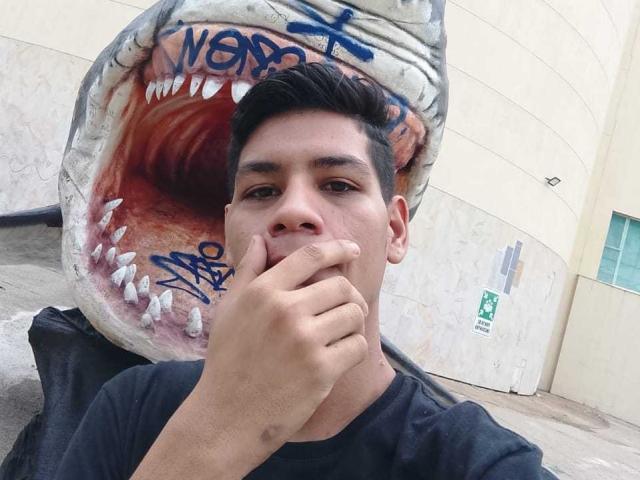 CristoferBlack with short black hair, dark shirt, covering mouth with hand, standing in front of giant shark mouth graffiti. Shark mouth has blue graffiti on white.