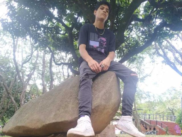 CristoferBlack sits on a large rock, wearing a black shirt, ripped jeans, white sneakers, and a necklace, looking up with a serious expression, surrounded by green trees.