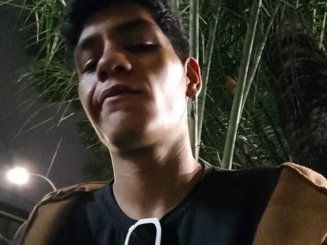 CristoferBlack has short black hair, a slight smile, and is wearing a black shirt with brown sleeves. His ear has a small hoop earring. Nighttime.