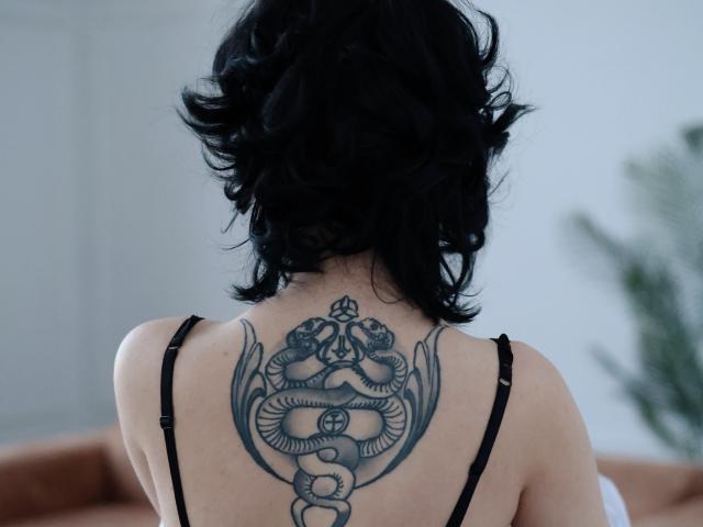 JulisSweet with black curly hair, back tattoo of a detailed snake and wings, wearing a black strap top, standing in a room.