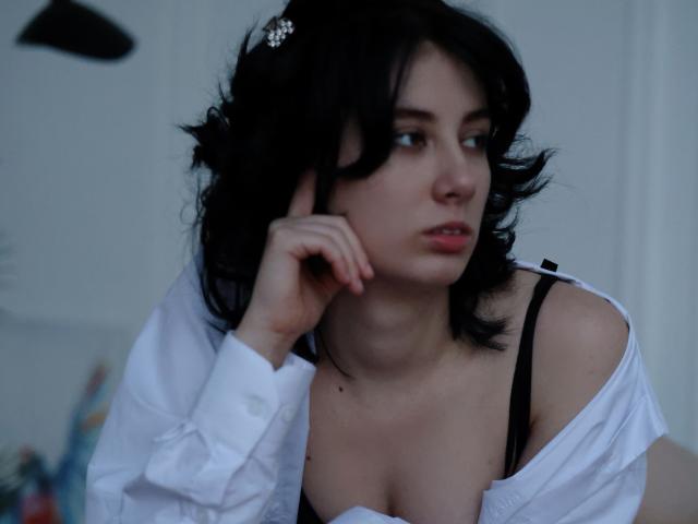 JulisSweet with black wavy hair, white shirt off shoulder, black bra, looking to the side, hand on cheek, pale skin, small jewelry on hair, soft blue light.