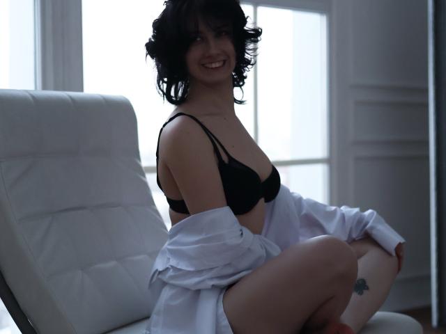 JulisSweet with short curly black hair, smiling, wearing a black bra and open white shirt, exposing her small tits and bare thighs, sitting on a white leather chair.