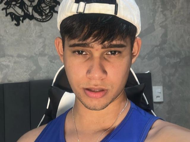 SmithWaylon with a goofy expression, wearing a white cap, blue tank top, and necklace, sitting in a black and white chair. His face is slightly pouty and he has dark hair.