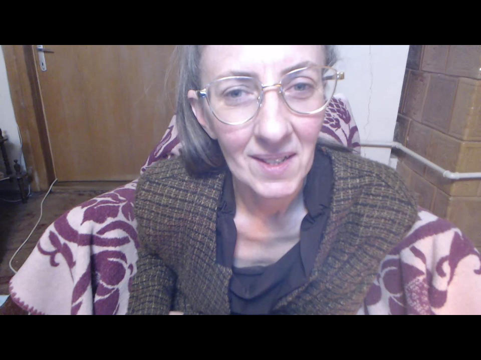 JoziAdoree, middle-aged with glasses, smiling, wearing a brown plaid sweater over a maroon top, has a slightly crooked nose and short brown hair, sitting in a wooden room.