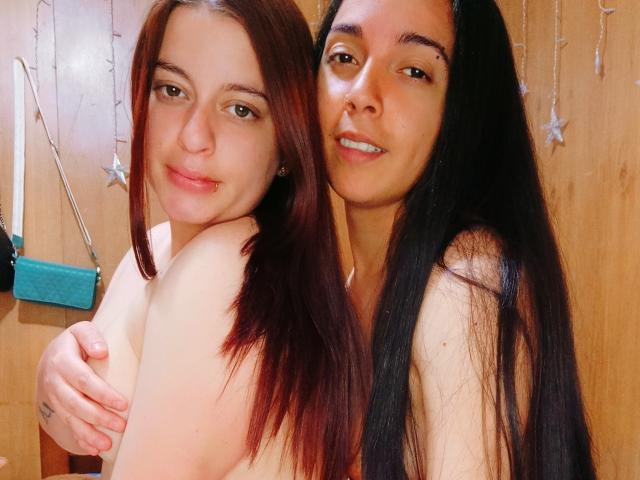 Two topless BarbarayLola69s with long red and black hair, holding each other, smiling at the camera. Boobs visible, one arm around each other's shoulders. Warm wooden background.
