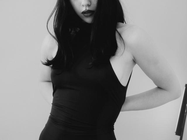Riverly, com batom preto, vestindo uma blusa preta justa sem mangas, expondo seus ombros e braços nus, em pé com as mãos atrás das costas, em uma fotografia em tons de cinza..