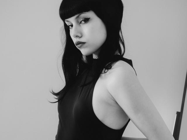 Riverly with dark bangs and black dress, showing side profile with raised arm, has pale skin, black eyeliner, and a serious expression. Her tits are visible through the dress.