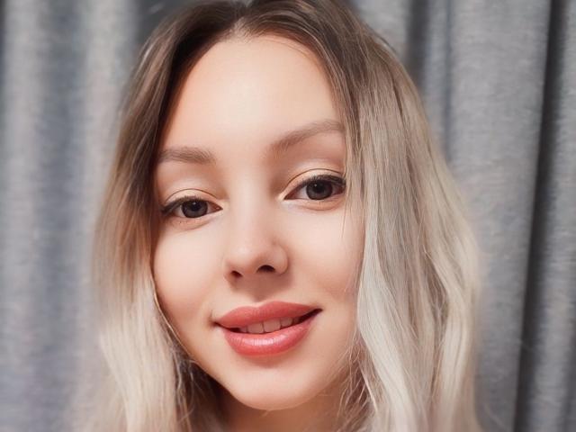 AliceSmiles has blonde, wavy hair, fair skin, and is smiling with pink lips. She has brown eyes and a slightly upturned nose. Her tits are not visible.