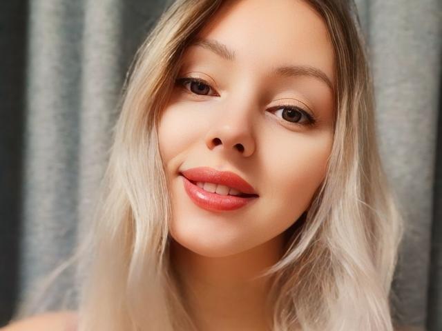 AliceSmiles has blonde hair, brown eyes, and shiny pink lips, smiling slightly with a cute, relaxed face. She's looking directly at the camera, with soft, natural makeup.