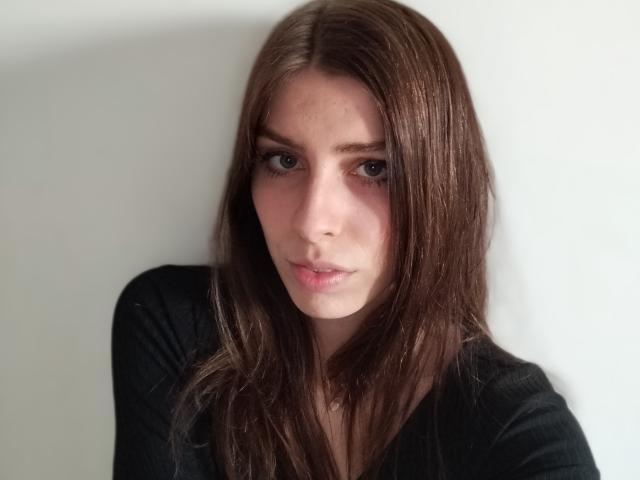 GlamPia has long brown hair, fair skin, and a serious expression, wearing a black top, with small, cute tits slightly visible. She's looking directly at the camera.