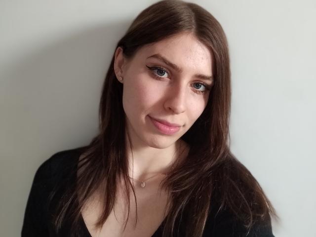 GlamPia has light skin, brown hair, and eyeliner, wearing a black top, with a subtle smile and a small necklace, looking at the camera.