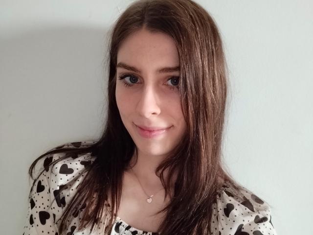 GlamPia has long brown hair, blue eyes, and a slight smile. She wears a heart-patterned shirt and a small heart necklace. Her tits are small and she's cute.