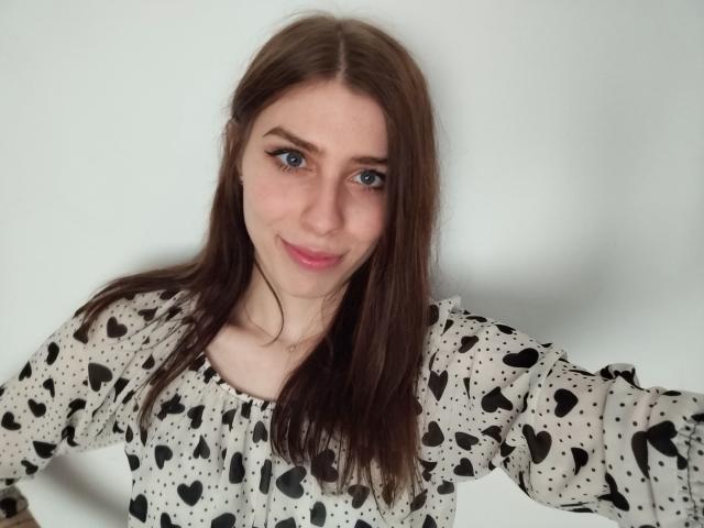 GlamPia has brown hair, blue eyes, and a small smile. She's wearing a black heart-patterned, long-sleeve shirt with a ruffled neckline. Her tits are small.