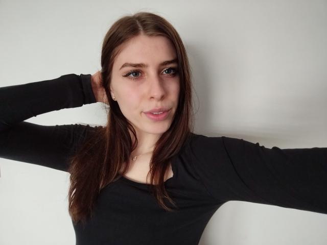 GlamPia with brown hair, black long-sleeve shirt, arms raised, looking at camera, slight smile, blue eyes, fair skin, small nose, subtle makeup, white background.