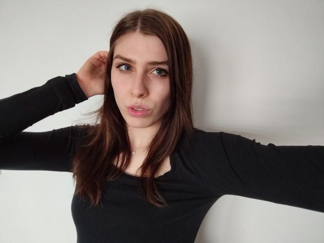 GlamPia has medium brown hair, blue eyes, and a serious expression, wearing a black long-sleeve shirt, with one hand behind her head, showing her small tits.