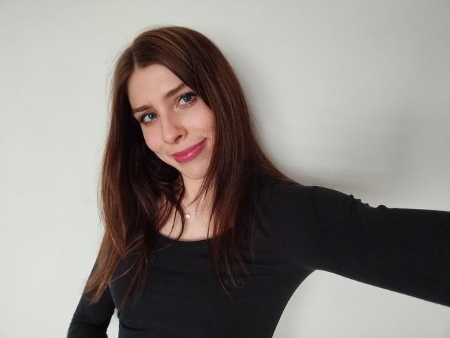 GlamPia with brown hair, pink lipstick, black long-sleeve shirt, small necklace, smiling slightly, taking a selfie, white background, right arm extended.