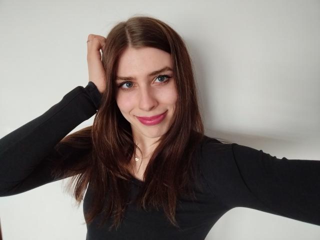 GlamPia with brown hair, blue eyes, pink lipstick, black long-sleeve shirt, right hand in hair, smiling, white background, subtle necklace, cute, attractive.