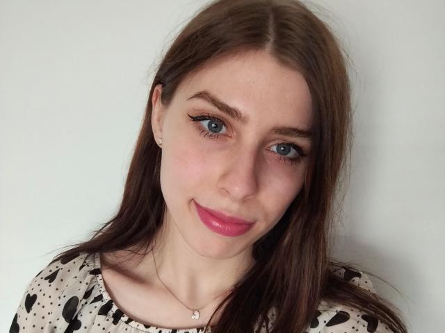 GlamPia with blue eyes, pink lipstick, and brown hair, wearing a heart-patterned top, smiling softly at the camera, necklace with small pendant, fair skin, cute face.