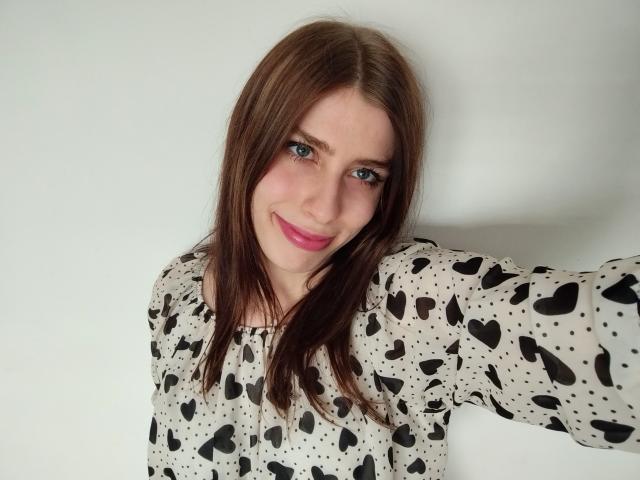 GlamPia has brown hair, blue eyes, pink lipstick, and a cute smile. She's wearing a black heart and dot patterned shirt, with her arm slightly raised.