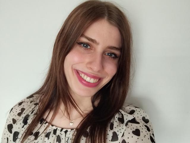 GlamPia has brown hair, blue eyes, pink lipstick, and a smile showing white teeth. She's wearing a black and white polka dot top with a small necklace.
