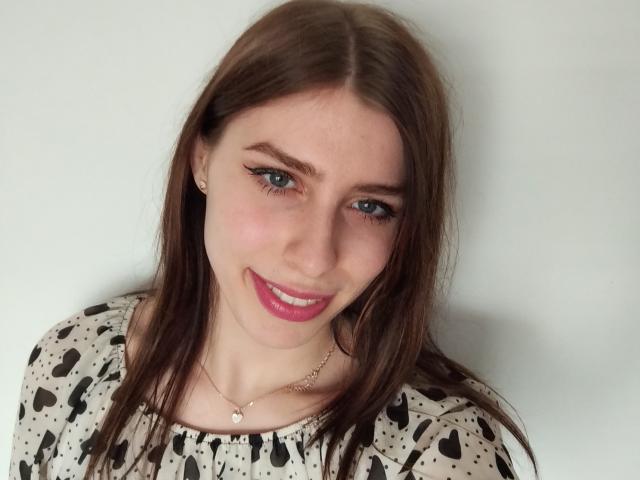 GlamPia has blue eyes, pink lipstick, and brown hair, wearing a black and white heart-patterned top, with a gold necklace and small earrings, smiling slightly.