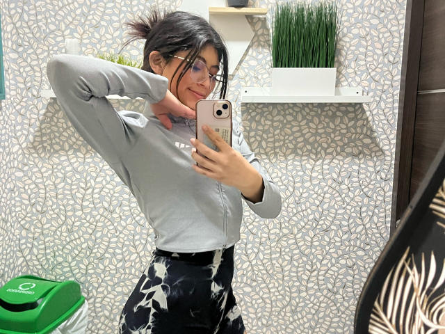 CoriinneBenson with black hair in a messy bun, wearing glasses, grey long-sleeve shirt, and black floral pants, taking a mirror selfie in a stylish room.