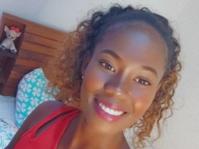 SandSexy tiene la piel de color marrón oscuro, el cabello rizado de color miel y está sonriendo mostrando sus dientes blancos, lleva una camiseta roja y se ve su rostro sin vello en un selfie..