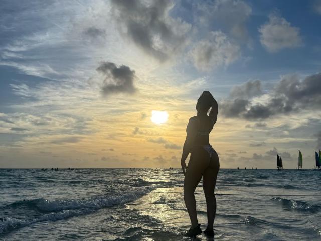 Heyly, a curvy woman with large breasts, stands in the ocean at sunset, silhouetted against the glowing sky, water splashing around her legs.