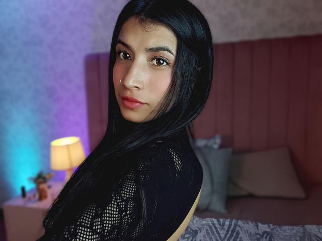 NaiaSanders with long black hair, wearing a black lace top, looking over her shoulder with a subtle smile, in a bedroom with a pink headboard and a lit lamp.
