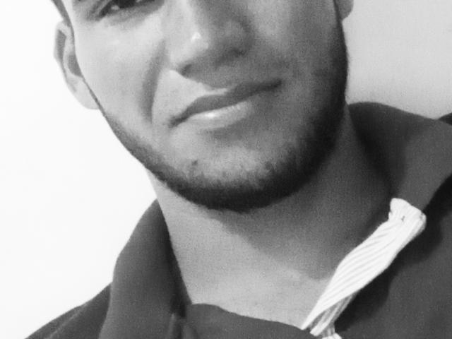 Croosty's face, slightly out of focus, with a short beard and a subtle smirk. He's wearing a dark shirt with a white tie, black and white image.