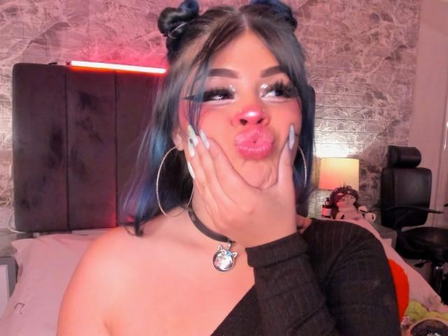 MissFontainne with blue hair in buns, wearing large hoop earrings and a black choker with a moon pendant, making a pouty face with her hands on her cheeks.
