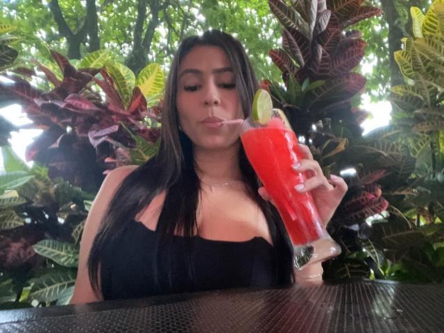 NahomyStone with long dark hair, black top, sipping a red cocktail with a lime, looking down. Boobs visible, holding drink in right hand, green leafy background.