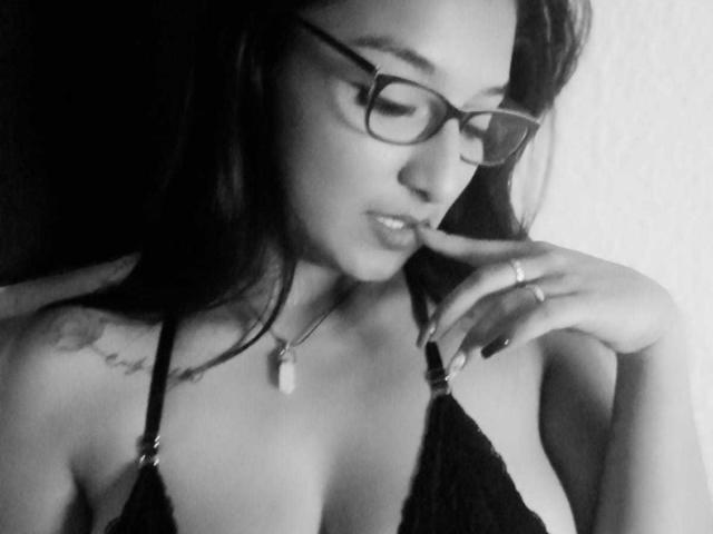 black and white photo of FantastykLadies with glasses, dark hair, ring on finger, black lace bra, visible tattoo on right shoulder, touching lips with finger, showing small tits.