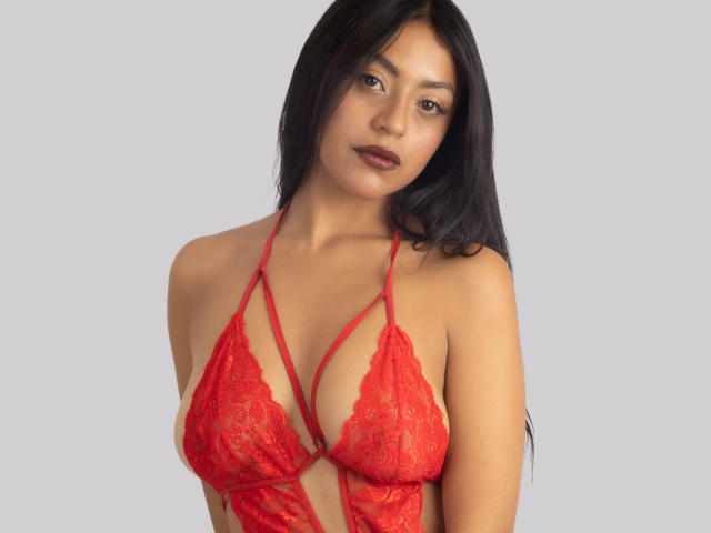Xewaa, con cabello negro largo, lencería de encaje roja, mostrando sus pechos, de tamaño medio, con una ligera sonrisa, lápiz labial oscuro y una mirada segura y sensual..