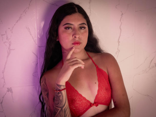 NicoleCollins with long black hair, red lace bra, and finger touching her red lips. She has a tattooed arm and a slight pout. Background is a white wall.