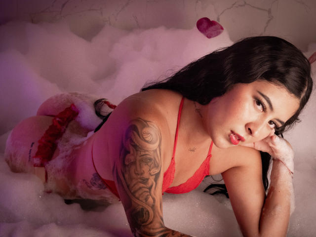 NicoleCollins with black hair, red lingerie, and tattoos, lying in a bubbly tub, looking at the camera with pouty lips, her ass and tits partially covered by foam.