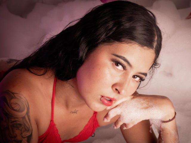 NicoleCollins with dark hair, red lace top, pouty lips, tattoo on left arm, soap on her cheek, in a bubble bath, looking seductively at the camera.
