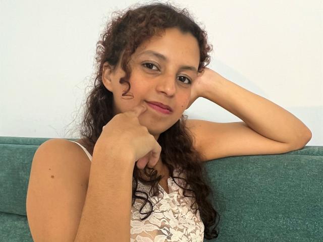 Emily Martin, avec ses cheveux bruns bouclés, sa peau brun clair et son rouge à lèvres rose, appuyant son menton sur sa main, portant un haut blanc en dentelle, assise sur un canapé vert..