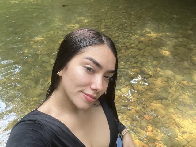 SharonKlart with wet dark brown hair, black shirt, and subtle smile, standing in clear water with visible rocks, looking at the camera with slightly parted lips.