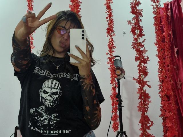 AdaWalton with tattoos, wearing a black skull shirt, making a peace sign, tongue out, holding a phone, red leaf garlands, and a microphone stand in the background.